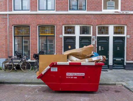A large red commercial waste skip positioned on a paved street in front of a row of traditional brick terraced houses with white window frames and dark green doors. The skip is filled with various types of rubbish, including flattened cardboard boxes, paper bags, and miscellaneous debris, occupying most of its open top. To the left of the skip, two bicycles are parked against the pavement, one with a black frame and the other with a brown frame, both secured with locks. The background features the brick façade of the buildings, with windows that have barred metal grilles and yellow curtains visible behind the glass. The scene appears to be part of a waste collection or rubbish removal operation in an inner-city residential area, with a focus on commercial waste disposal services provided by Commercial Waste Ascot operating in the local vicinity. The sky is overcast, and the street surface is clean, with no other visible vehicles or pedestrians in the image.