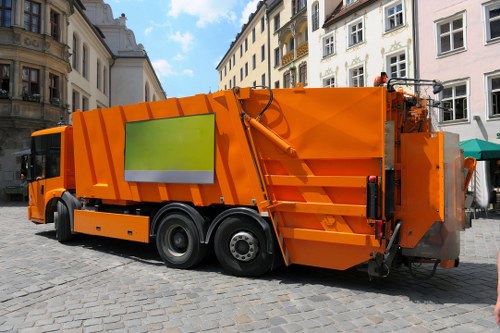 A large, modern orange rubbish collection vehicle parked on a cobbled street in an urban area. The truck features a prominent, reflective lime-green panel on its side, and its body is made of metal with a textured, ribbed finish. The vehicle is equipped with hydraulic mechanisms at the rear for waste compaction and collection. It is positioned in front of Georgian-style buildings with white and beige facades, multiple windows, and decorative architectural details, typical of a historic town centre. The scene appears to be during daylight hours with natural lighting, and there are no visible waste materials or bins in the image. The vehicle belongs to Commercial Waste Ascot, serving the local area around Ascot and surrounding towns, supporting rubbish removal services with professional, city-based waste management solutions.