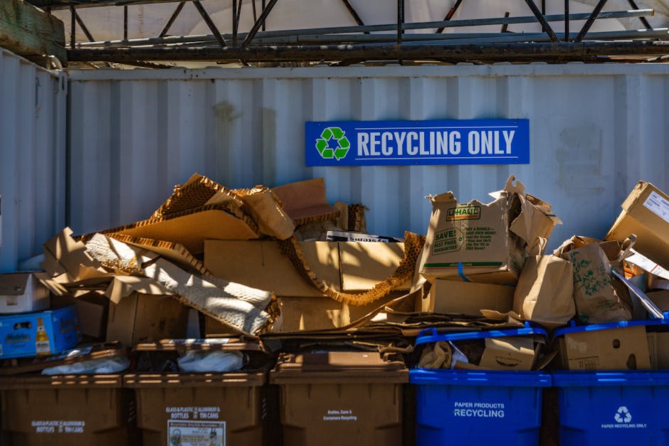 A collection of various flattened cardboard boxes and paper packaging materials stacked on top of black and blue recycling bins, situated against a grey metal container wall with a large blue sign tha